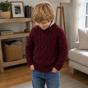 Può includere: Un maglione a maniche lunghe in maglia a trecce color bordeaux intenso, caratterizzato da un motivo testurizzato a rombi e corde sul davanti. Il maglione presenta uno scollo rotondo ed è indossato sopra una camicia con un motivo a quadri.