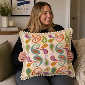 May include: A square throw cushion with a cream-coloured base featuring a repeating pattern of colourful paisley shapes in shades of orange, purple, green, and blue. The edges of the cushion are finished with a decorative cream-coloured pom-pom trim.