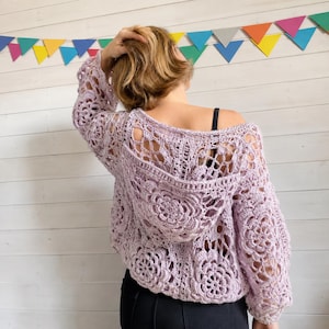 Puede incluir: Un suéter de ganchillo de manga larga en color violeta claro con un intrincado patrón de motivos florales en toda la prenda. El suéter incluye una capucha y un diseño de punto abierto con un escote ancho, mostrando una textura detallada.