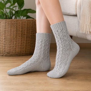 Könnte beinhalten: Ein Paar hellgraue Strickstrümpfe mit einem vertikalen Zopfmuster in Kombination mit einem komplizierten Lochmuster. Die Socken verfügen über ein geripptes Bündchen am oberen Rand, einen verstärkten Fersenbereich und eine glatte Zehenpartie.