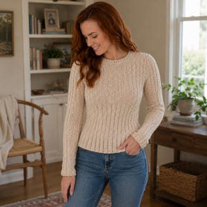 May include: A long-sleeved, cream-colored knit sweater featuring a ribbed texture throughout the body and sleeves, a round neckline, and a fitted silhouette. The sweater is paired with blue denim jeans, creating a casual and neutral ensemble.