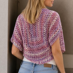 Può includere: Un top all'uncinetto a maniche corte con un motivo di filato variegato nei toni del rosa, viola e bianco. Il capo presenta una lavorazione a maglia aperta e ariosa con un orlo smerlato, indossato sopra una base bianca e jeans blu.