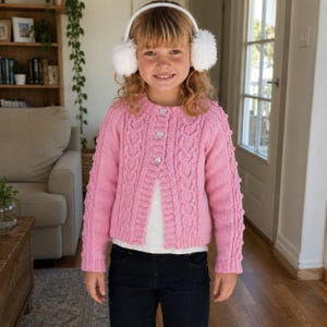 May include: A bright pink cable-knit cardigan featuring a round neckline, long sleeves, and three heart-shaped buttons down the front. The sweater is worn over a plain white shirt and paired with dark blue jeans, complemented by white earmuffs.