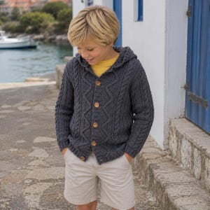 Può includere: Un cardigan con cappuccio in maglia a trecce grigio scuro con chiusura frontale a cinque bottoni in legno marrone chiaro, indossato sopra una t-shirt gialla a tinta unita e pantaloncini beige chiaro, con un motivo a maglia testurizzato.