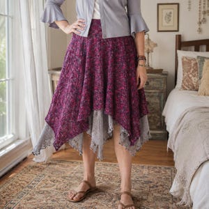 May include: A layered midi skirt featuring a deep magenta and purple textured top layer with an asymmetrical handkerchief hemline, worn over a light gray lace-trimmed underskirt. The skirt is paired with a light gray long-sleeved top.