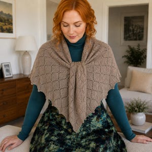 May include: A triangular knit shawl in a light brown color featuring a repeating diamond lace pattern with a scalloped edge. The shawl is draped over a dark teal long-sleeved top and a dark green and black patterned skirt.