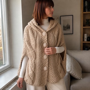 May include: A beige hooded poncho crafted from thick knit yarn featuring a cable knit pattern and four large, round, fabric-covered buttons down the front. The garment has wide arm openings and a textured surface, worn over a white long-sleeve shirt.