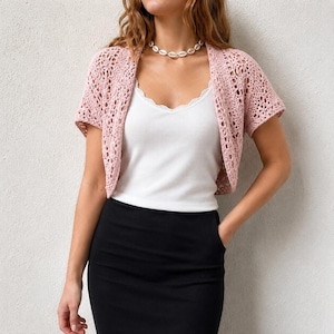 May include: A light pink, short-sleeved, open-front crochet bolero cardigan featuring an intricate, airy diamond-patterned knit. The garment is worn over a white, ribbed, sleeveless camisole with a delicate lace trim along the neckline.