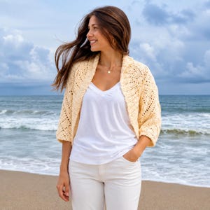 May include: A pale yellow, open-front crochet shrug featuring a textured, diamond-patterned knit design with three-quarter length sleeves, worn over a plain white V-neck t-shirt and white trousers.