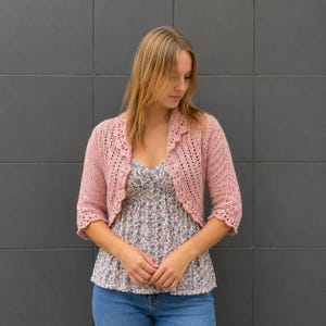Op de afbeelding: Een oudroze gebreid bolero-vest met een open voorzijde, driekwartmouwen en een geschulpte rand. Het kledingstuk wordt gedragen over een top met bloemenprint, een hartvormige halslijn en een blauwe spijkerbroek, met een gestructureerd breipatroon.