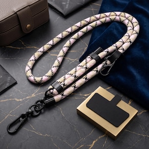 May include: A long, cylindrical rope strap featuring a repeating houndstooth pattern in shades of light pink, beige, and black. The strap includes black metal hardware, including two swivel lobster clasps and two black leather-like connectors.