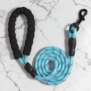 May include: A bright blue rope lead featuring a black foam-padded handle and a black metal swivel clasp. The lead is constructed from a durable braided cord with reflective black threading woven throughout the material for visibility.
