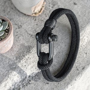 Puede incluir: Pulsera de cuerda negra con cierre negro mate. La pulsera está hecha de un material grueso y tejido y presenta un cierre en forma de U. La pulsera se muestra sobre una superficie gris con una pequeña planta en maceta en el fondo.