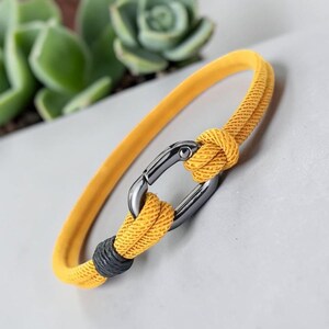 Può includere: Un bracciale in corda gialla con una chiusura a moschettone ovale in metallo scuro. La corda presenta una trama testurizzata e include un piccolo nodo decorativo grigio scuro vicino alla chiusura, che unisce le estremità della corda.