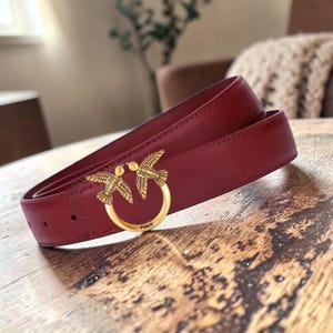 May include: A deep red leather belt featuring a circular gold-tone metal buckle with two symmetrical bird figures facing each other. The buckle is engraved with the word PINKO at the bottom center. The belt strap has visible stitching along the edges.