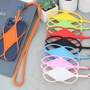 Puede incluir: Un smartphone azul oscuro asegurado en una funda de silicona naranja con cordón para colgar. Al lado, una pila de soportes de silicona idénticos en amarillo, verde, rojo, negro, azul claro, rosa y blanco, cada uno con una correa larga a juego.