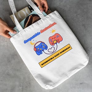 May include: A white canvas tote bag featuring a graphic of two cartoon game controllers connected by a heart-shaped cord. The blue controller wears headphones under a crescent moon, while the red controller holds a mug under a sun. Text reads: Compatible Controllers, INCOMPATIBLE SLEEP SCHEDULES.