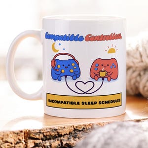 May include: A white ceramic mug featuring a blue gaming controller with headphones under a moon and a red gaming controller with a coffee mug under a sun. The text reads Compatible Controllers, and below, INCOMPATIBLE SLEEP SCHEDULES in a yellow box.