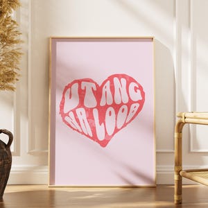 Può includere: Una stampa artistica rosa incorniciata con un cuore rosso e le parole "UTANG NA LOOB" in bianco. L'opera d'arte è esposta su un pavimento in legno, accanto a un vaso decorativo con piante essiccate.