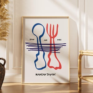 May include: A framed art print featuring a blue spoon and a red fork design. The words "Spoon and Fork" are above the utensils, with "Mancan Tayon!" below. The print is in a gold frame, leaning against a white wall.