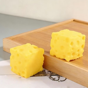 May include: Two yellow, cube-shaped objects with holes, resembling cheese, are displayed on a wooden tray. The objects have a textured surface and are brightly colored. The tray is made of light-colored wood.