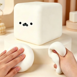 Op de afbeelding: Een wit, kubusvormig nachtlampje met een cartoon zeehondengezicht. Het licht staat op een witte sokkel, met een witte, ronde stressbal en een witte, langwerpige stressbal die in de handen wordt gehouden.