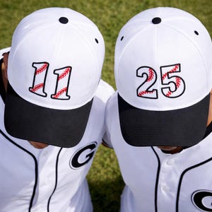 Puede incluir: Dos gorras de béisbol blancas con viseras negras. Una gorra muestra el número "11" en rojo con costuras de béisbol, la otra muestra "25". Ambas gorras tienen un botón negro en la parte superior.