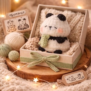 May include: A handmade crochet panda bear with black and white fur, a green scarf, and pink cheeks, presented in a gift box with a green bow. The box sits on a wooden tray with a soft, textured blanket and string lights.