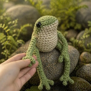 May include: A handmade crochet frog in shades of green and beige sits on a rock. The frog has black eyes and long, slender legs. The frog is in a natural outdoor setting, with water and greenery in the background.