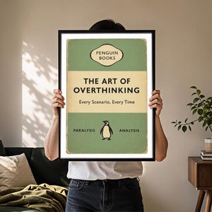 May include: A framed rectangular wall art print featuring a vintage-style book cover design in sage green and cream. The text reads PENGUIN BOOKS, THE ART OF OVERTHINKING, Every Scenario, Every Time, and PARALYSIS ANALYSIS, with a small penguin illustration.