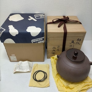 以下が含まれることがあります： 茶色の蓋付きの陶器のティーポットが、黄色の布の上に置かれ、茶色のリボンで結ばれた木箱の隣にあります。 青と白の模様の蓋が付いた段ボール箱もあります。