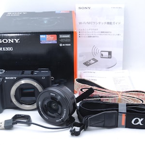 May include: A black Sony α6300 mirrorless digital camera kit with a lens, strap, and box. The box is black with white text and a red accent. The kit includes a USB cable and instruction manuals.