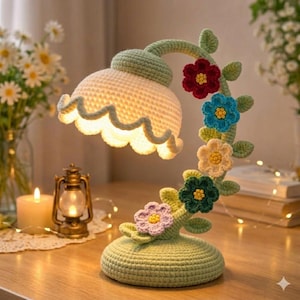 May include: A handcrafted crochet lamp in the shape of a flower. The lamp features a light green base and stem, with a white scalloped lampshade. Colorful crochet flowers in red, blue, yellow, green, and purple adorn the stem. The lamp is a decorative home accent.