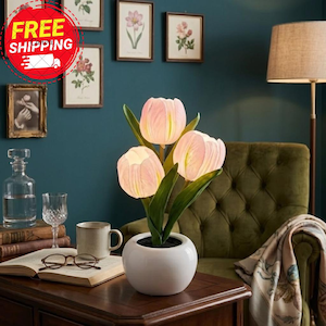 May include: A white ceramic pot with three illuminated pink tulip-shaped lights. The tulips have green stems and leaves. The lamp is on a wooden table, with a mug, glasses, and a book nearby. The background includes framed botanical prints.