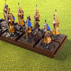 May include: Miniature figurines of archers in yellow and blue attire, each holding a bow and arrow. The figures are mounted on individual square bases, arranged on a rectangular brown base, and set against a green backdrop.