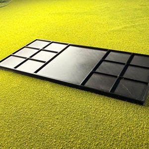 May include: A black rectangular tray with several compartments. The tray is divided into sections with raised edges, forming a grid pattern. The surface has a textured look, and the tray is set against a bright green background.