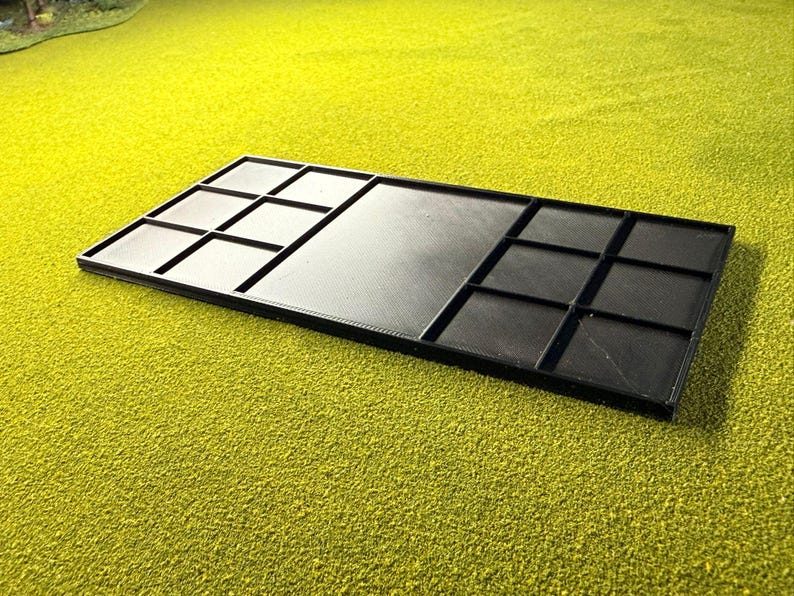 May include: A black rectangular tray with several compartments. The tray is divided into sections with raised edges, forming a grid pattern. The surface has a textured look, and the tray is set against a bright green background.