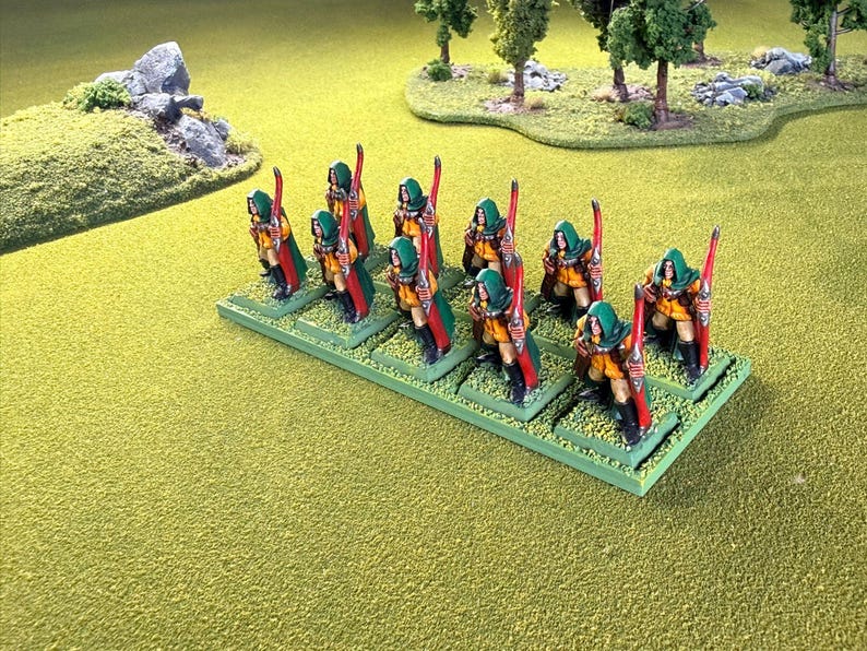 Op de afbeelding: Miniatuurbeeldjes van boogschutters in groene capuchonmantels en gele tunieken, elk met een rode boog. De figuren zijn gemonteerd op groene bases, tegen een grasachtig terrein met miniatuurbomen en rotsen.