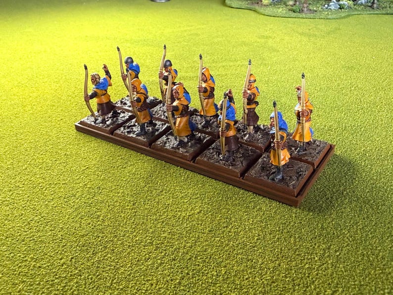 May include: Miniature figurines of archers in yellow and blue attire, each holding a bow and arrow. The figures are mounted on individual square bases, arranged on a rectangular brown base, and set against a green backdrop.