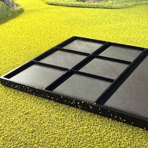 May include: A black, rectangular tray with multiple compartments. The tray is divided into sections by raised dividers, creating separate spaces. The surface of the tray has a textured pattern. The tray is set against a bright green, textured background.