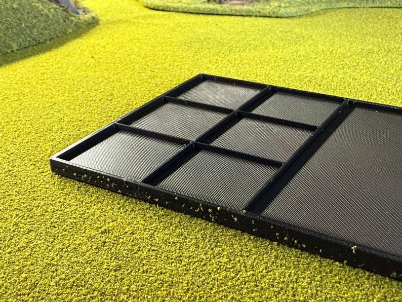 May include: A black, rectangular tray with multiple compartments. The tray is divided into sections by raised dividers, creating separate spaces. The surface of the tray has a textured pattern. The tray is set against a bright green, textured background.