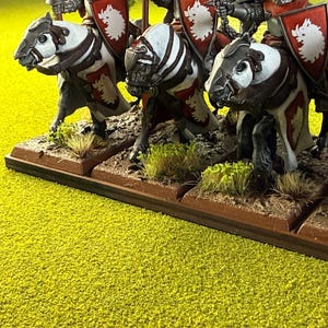 Peut inclure: Figurines miniatures de chevaliers &agrave; cheval, avec des chevaux blancs et bruns. Les chevaliers portent des boucliers rouges avec un embl&egrave;me blanc. Les figurines sont sur une base marron avec un terrain vert et marron, sur un fond jaune-vert.
