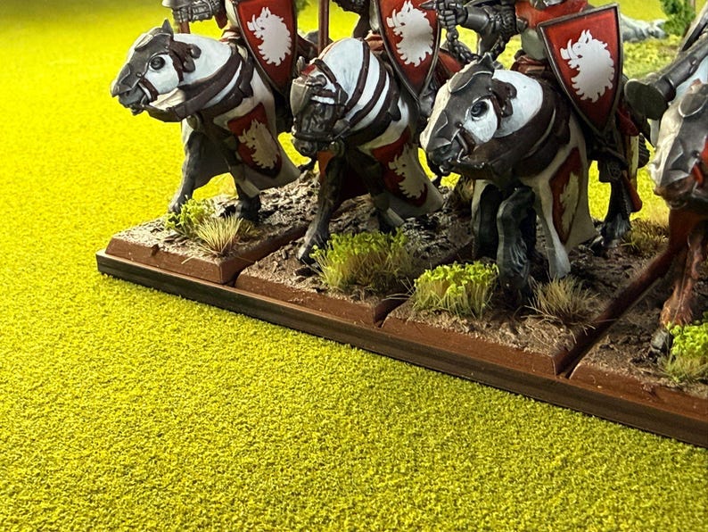 Peut inclure: Figurines miniatures de chevaliers &agrave; cheval, avec des chevaux blancs et bruns. Les chevaliers portent des boucliers rouges avec un embl&egrave;me blanc. Les figurines sont sur une base marron avec un terrain vert et marron, sur un fond jaune-vert.