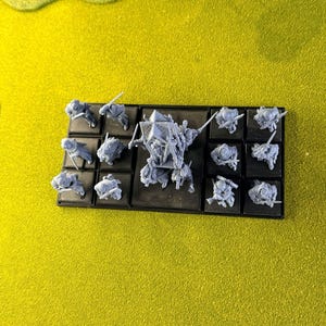 May include: Miniature grey figurines, possibly for a tabletop game, displayed on a black base. The figures represent soldiers and a larger central character. The background is a bright yellow.