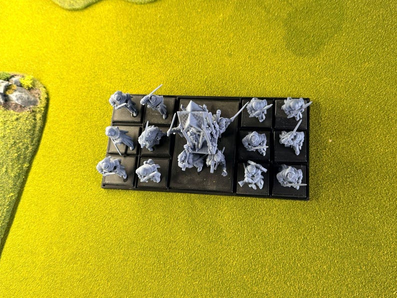 May include: Miniature grey figurines, possibly for a tabletop game, displayed on a black base. The figures represent soldiers and a larger central character. The background is a bright yellow.