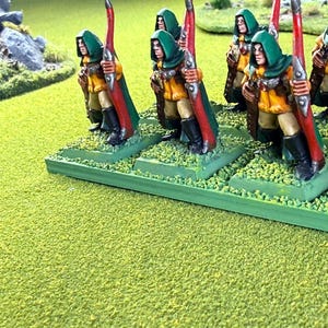 Op de afbeelding: Miniatuurfiguren van boogschutters in groene capuchonmantels, gele tunieken en rode bogen. De figuren staan op groene bases met getextureerd terrein, gerangschikt op een groen oppervlak. De sc&egrave;ne roept een fantasie- of historisch thema op.