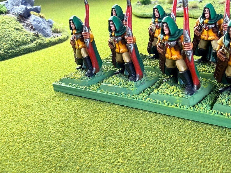 Op de afbeelding: Miniatuurfiguren van boogschutters in groene capuchonmantels, gele tunieken en rode bogen. De figuren staan op groene bases met getextureerd terrein, gerangschikt op een groen oppervlak. De sc&egrave;ne roept een fantasie- of historisch thema op.