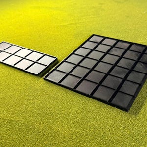 May include: Two black plastic trays with a grid pattern are shown on a green surface. One tray is rectangular with 12 squares, and the other is larger with 48 squares. The trays have a textured surface.