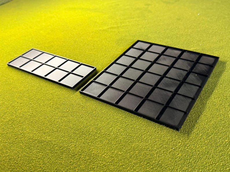 May include: Two black plastic trays with a grid pattern are shown on a green surface. One tray is rectangular with 12 squares, and the other is larger with 48 squares. The trays have a textured surface.