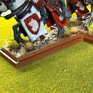 Peut inclure: Figurines miniatures de chevaliers &agrave; cheval. Les chevaux sont blancs et noirs, avec une armure d&eacute;taill&eacute;e et des &eacute;cussons h&eacute;raldiques rouges et blancs. La base est marron avec de l'herbe et de la terre factices, pos&eacute;e sur une surface verte.
